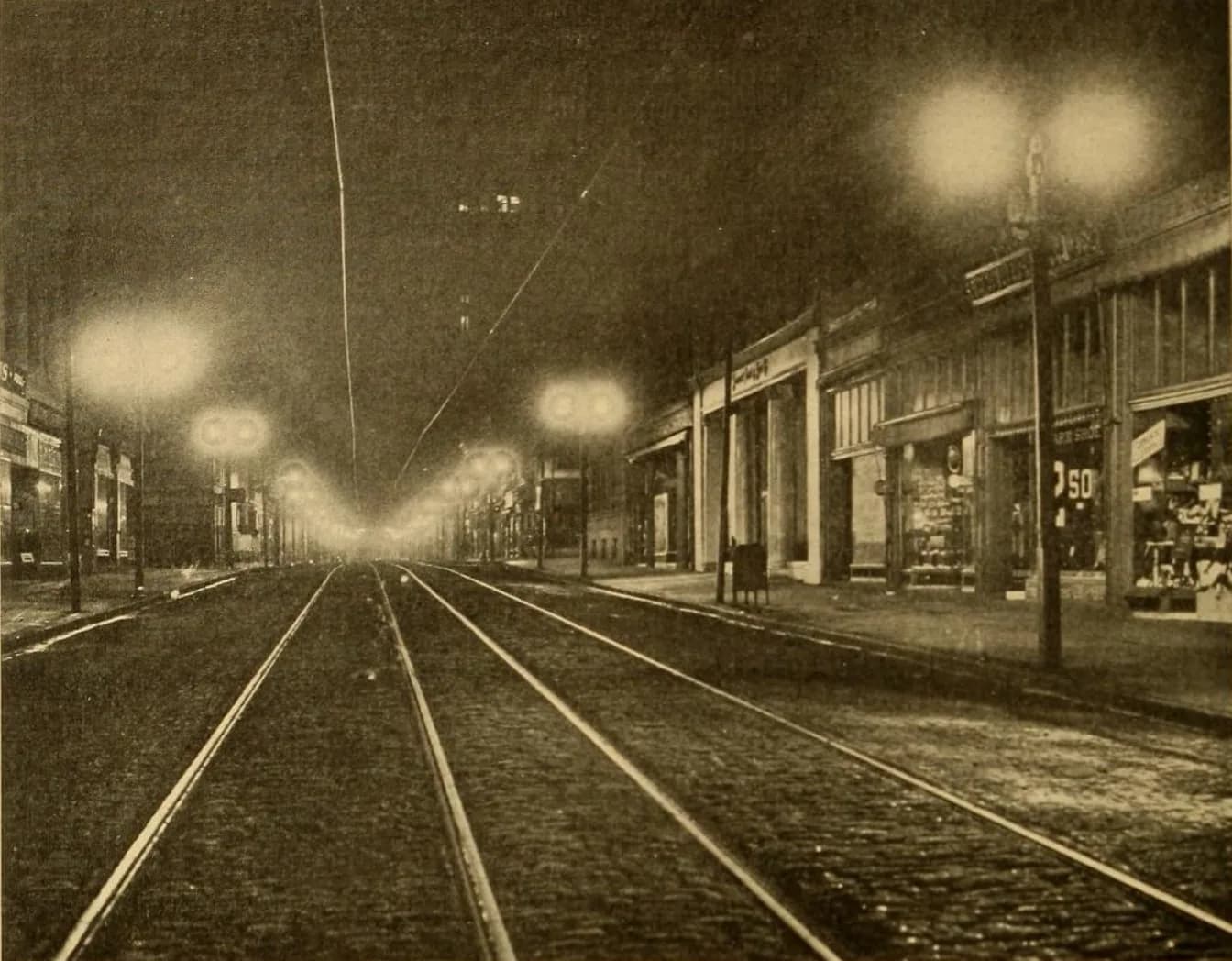 Sepia photograph of a city street at night lit by several very bright glowing lamps along tram tracks, with storefronts on the right.