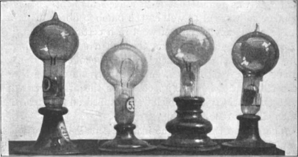 Sepia photograph of four vintage light bulbs of different shapes mounted upright on wooden bases, side by side.