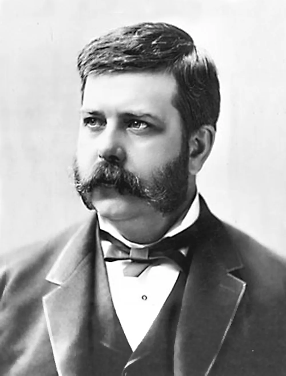 Black-and-white studio portrait of a mustached man in a dark suit and bow tie, looking to the side.