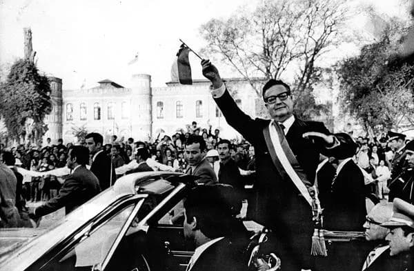A black-and-white photo of a man in a sash waving from an open convertible car to a large crowd in front of a historic building.