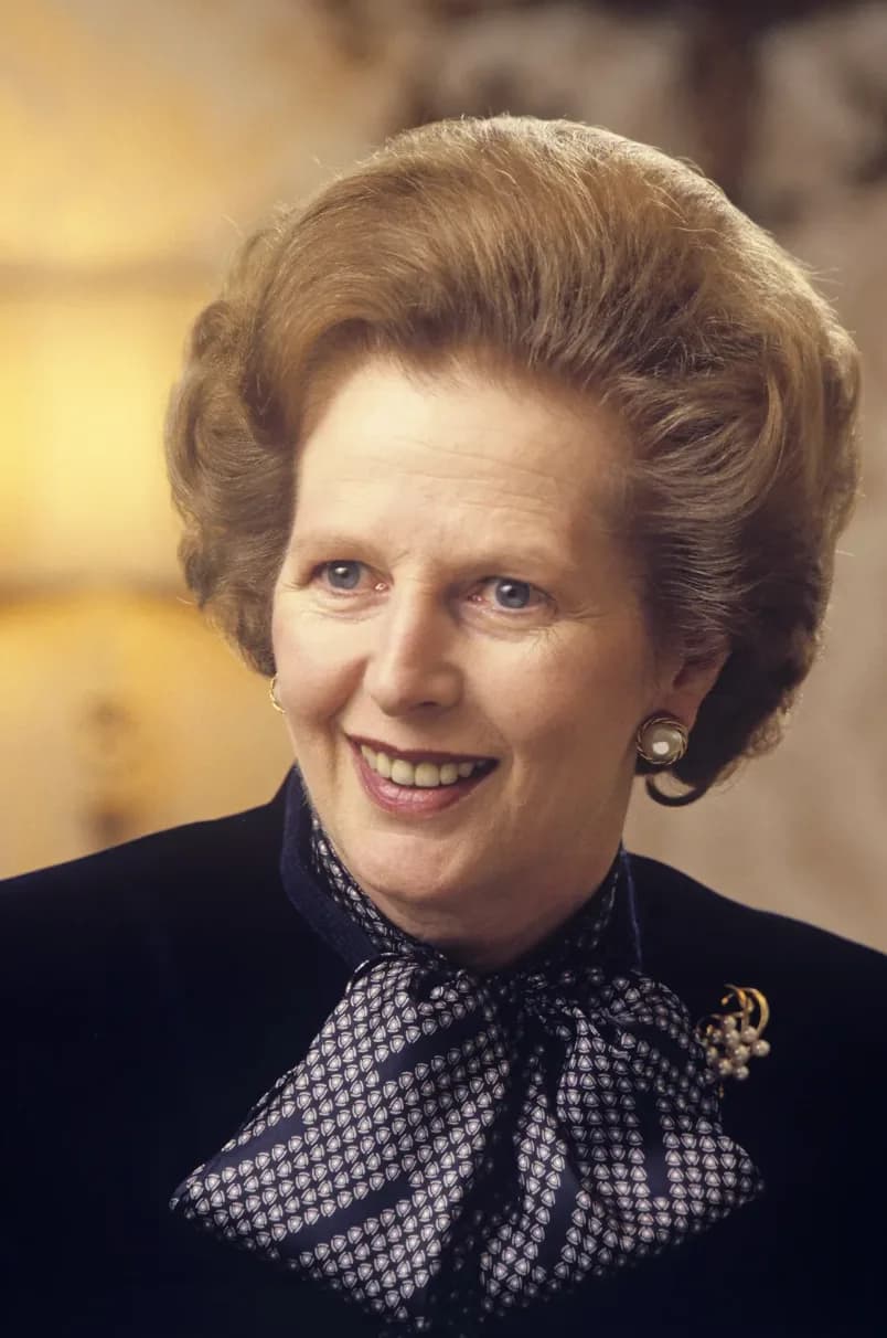 Color studio portrait of Margaret Thatcher smiling, wearing a dark blazer with a patterned bow blouse and pearl earrings.